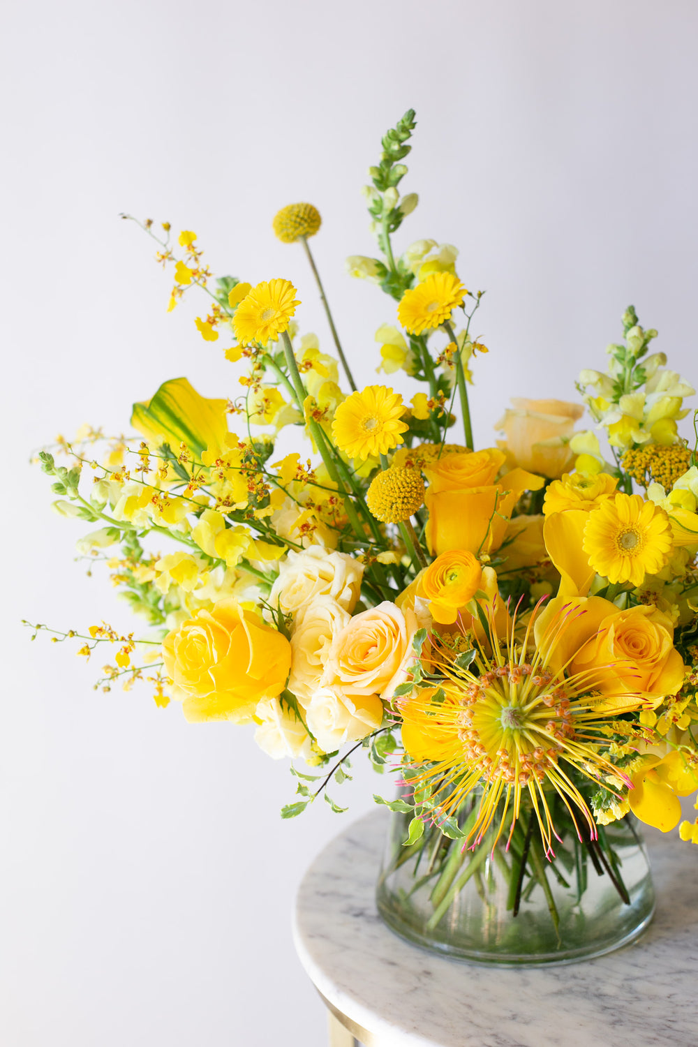 A flower arrangement in a glass container in front of a white backdrop on a small marble table. It's filled with yellow and gold flowers, including rose, billy balls, spray rose, yarrow, snap dragon, calla lily, and oncidium orchid. 