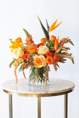 A flower arrangement in a glass container in front of a white backdrop on a small table. It's filled with orange and tangerine-colored flowers, including rose, snapdragon, ranunculus, bird of paradise, amaranthus, orchid, and pincushion protea.