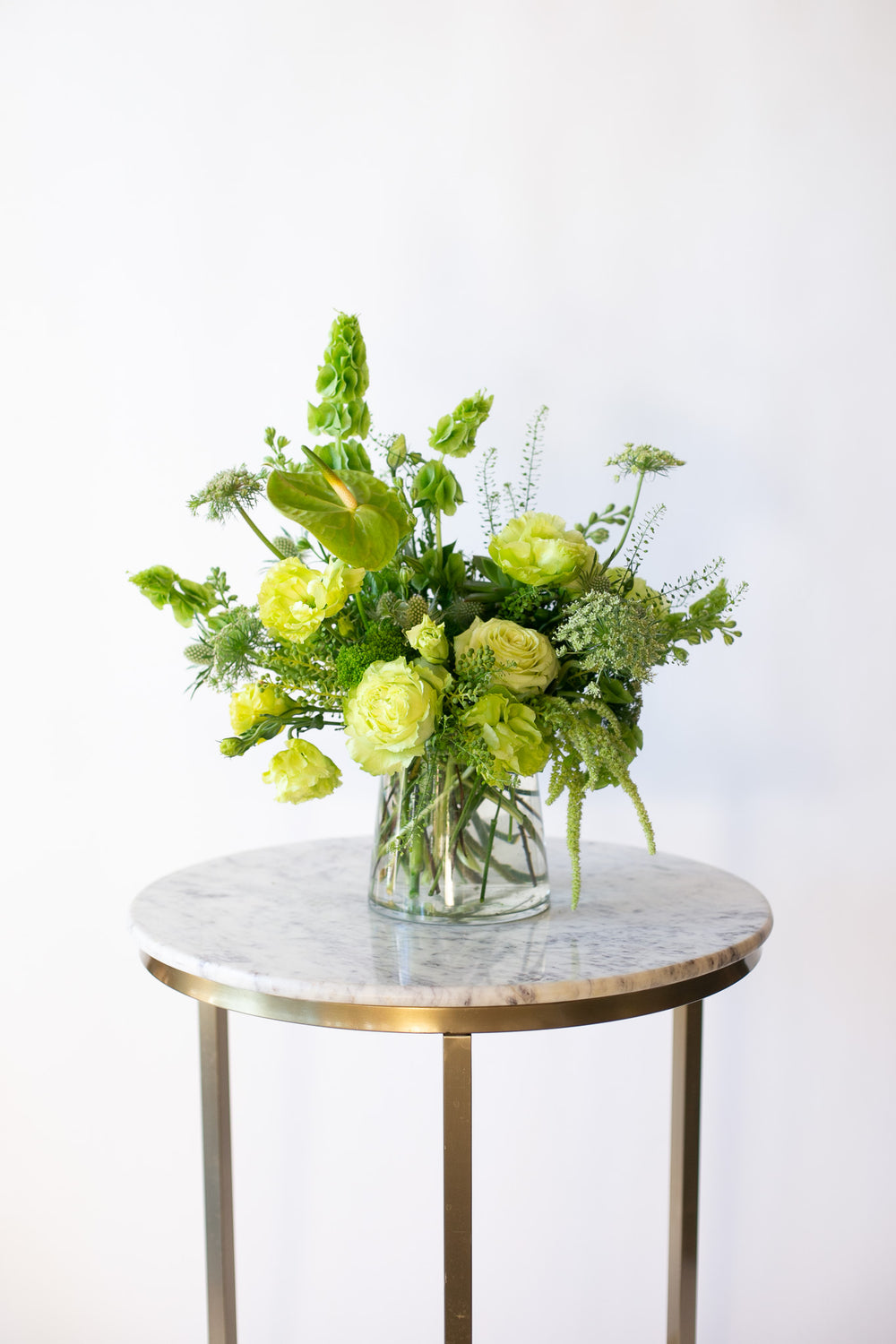 A flower arrangement in a glass container in front of a white backdrop on a small marble table. It's filled with green and chartreuse flowers and foliage, including rose, lisianthus, bells of Ireland, anthurium, Queen Anne's lace, and amaranthus. 