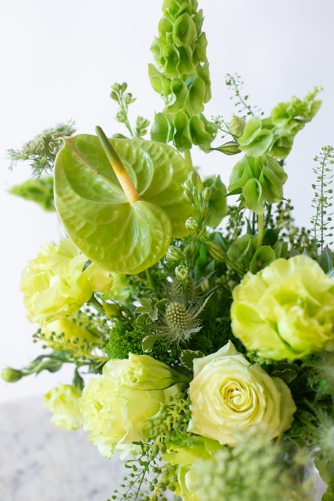 Closeup of a flower arrangement in a glass container in front of a white backdrop on a small marble table. It's filled with green and chartreuse flowers and foliage, including rose, lisianthus, bells of Ireland, anthurium, Queen Anne's lace, and amaranthus. 