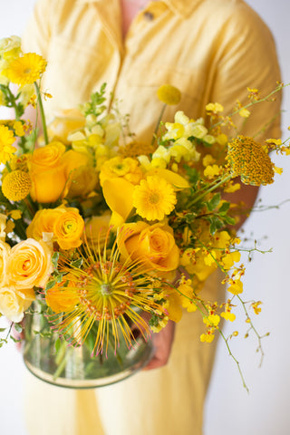 Woman in yellow holding A flower arrangement in a glass container in front of a white backdrop. It's filled with yellow and gold flowers, including rose, billy balls, spray rose, yarrow, calla lily, and oncidium orchid. 