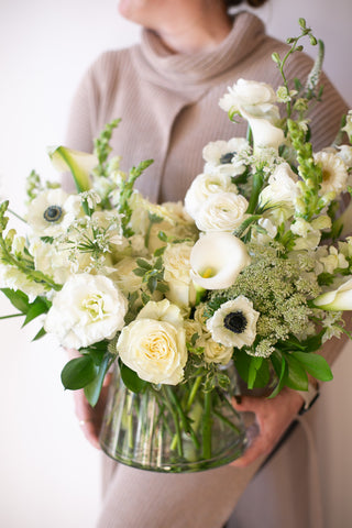 a woman in a taupe sweater holding A flower arrangement in a glass container in front of a white backdrop. It's filled with white flowers, including rose, lisianthus, Queen Anne's lace, calla lily, veronica, snapdragon, and anemone. 