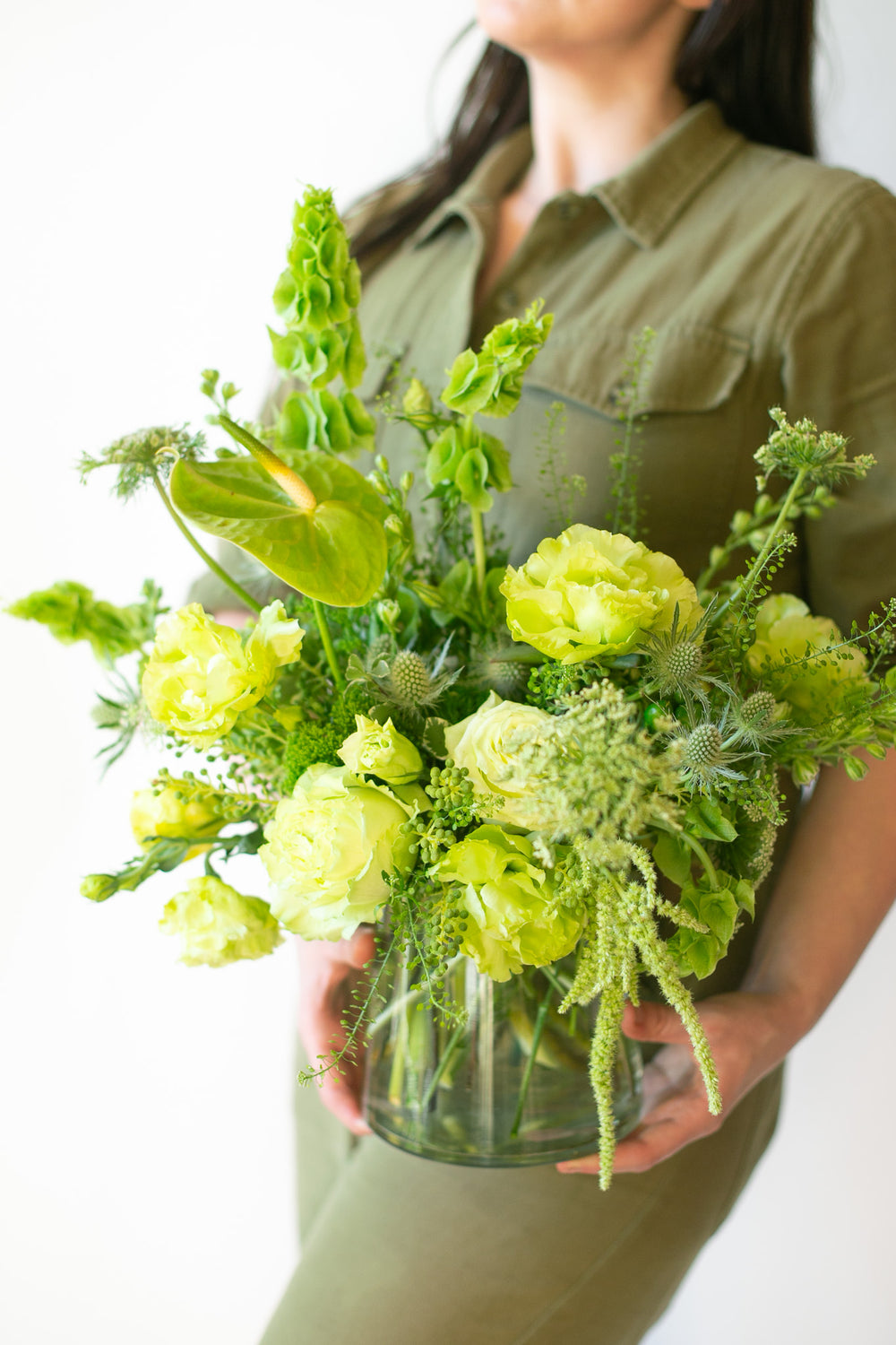 Woman in green jumpsuit holding a flower arrangement in a glass container in front of a white backdrop. It's filled with green and chartreuse flowers and foliage, including rose, lisianthus, bells of Ireland, anthurium, Queen Anne's lace, and amaranthus. 