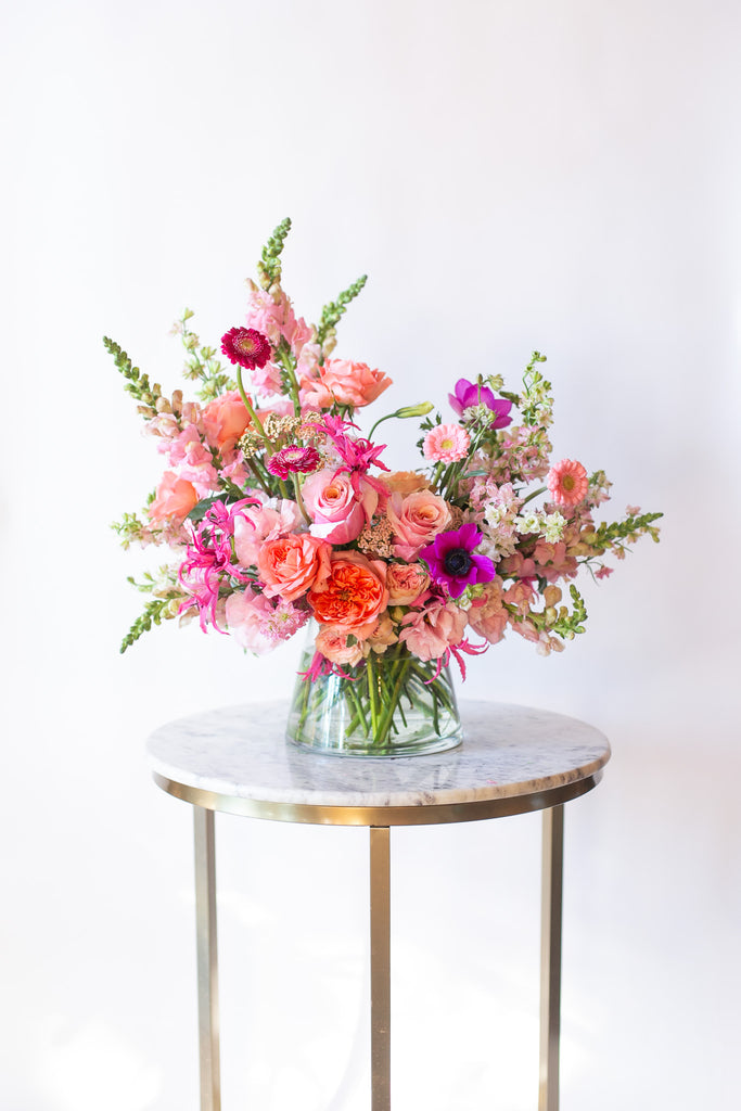 A flower arrangement in a glass container in front of a white backdrop on a small marble table. It's filled with pink, fuchsia, and magenta flowers, including rose, piccolini daisy, noreen lily, snapdragon, and delphinium. 