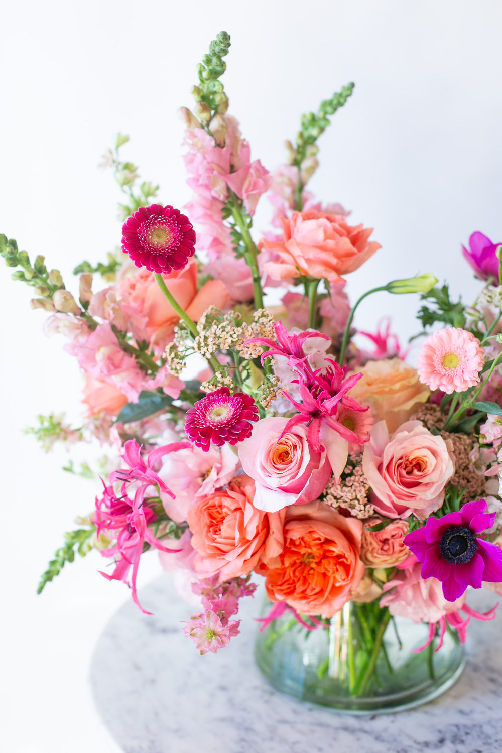 closeup of A flower arrangement in a glass container in front of a white backdrop on a small marble table. It's filled with pink, fuchsia, and magenta flowers, including rose, piccolini daisy, noreen lily, snapdragon, and delphinium. 