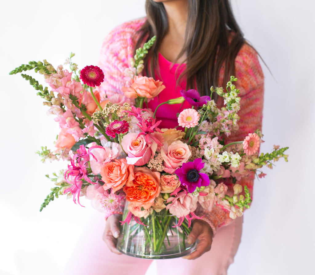 Woman in pink holding A flower arrangement in a glass container in front of a white backdrop on a small marble table. It's filled with pink, fuchsia, and magenta flowers, including rose, piccolini daisy, noreen lily, snapdragon, and delphinium. 