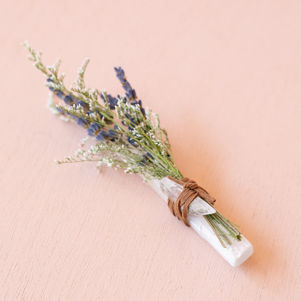 A bundled product flat lay featuring a small dried floral bouquet tied to a stick of selenite and a small quartz crystal on a against a pink background.
