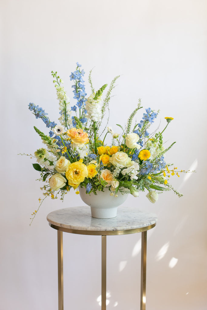 large WildFlora arrangement with a blend of yellow, blue, and white florals ina large white compote vase.