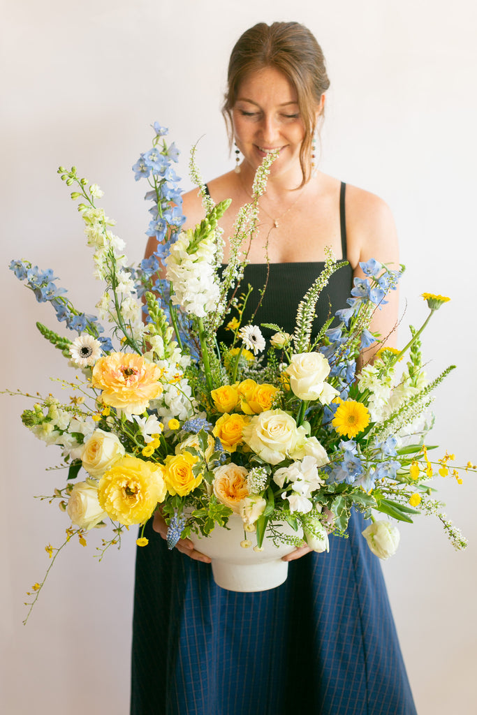 woman holding a large WildFlora arrangement with a blend of yellow, blue, and white florals in a large white compote vase.