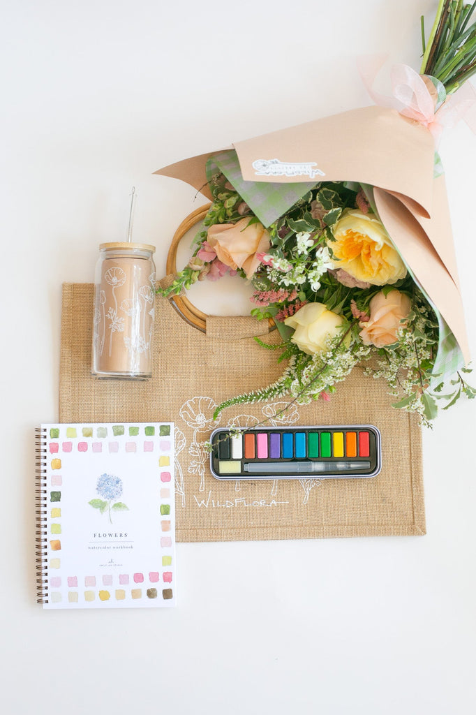 Bouquet of flowers on a jute tote with watercolors, a tumbler, and a painting book on a white surface