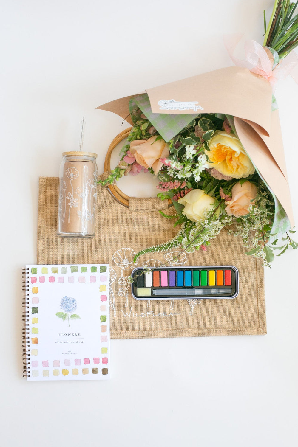 Bouquet of flowers on a jute tote with watercolors, a tumbler, and a painting book on a white surface