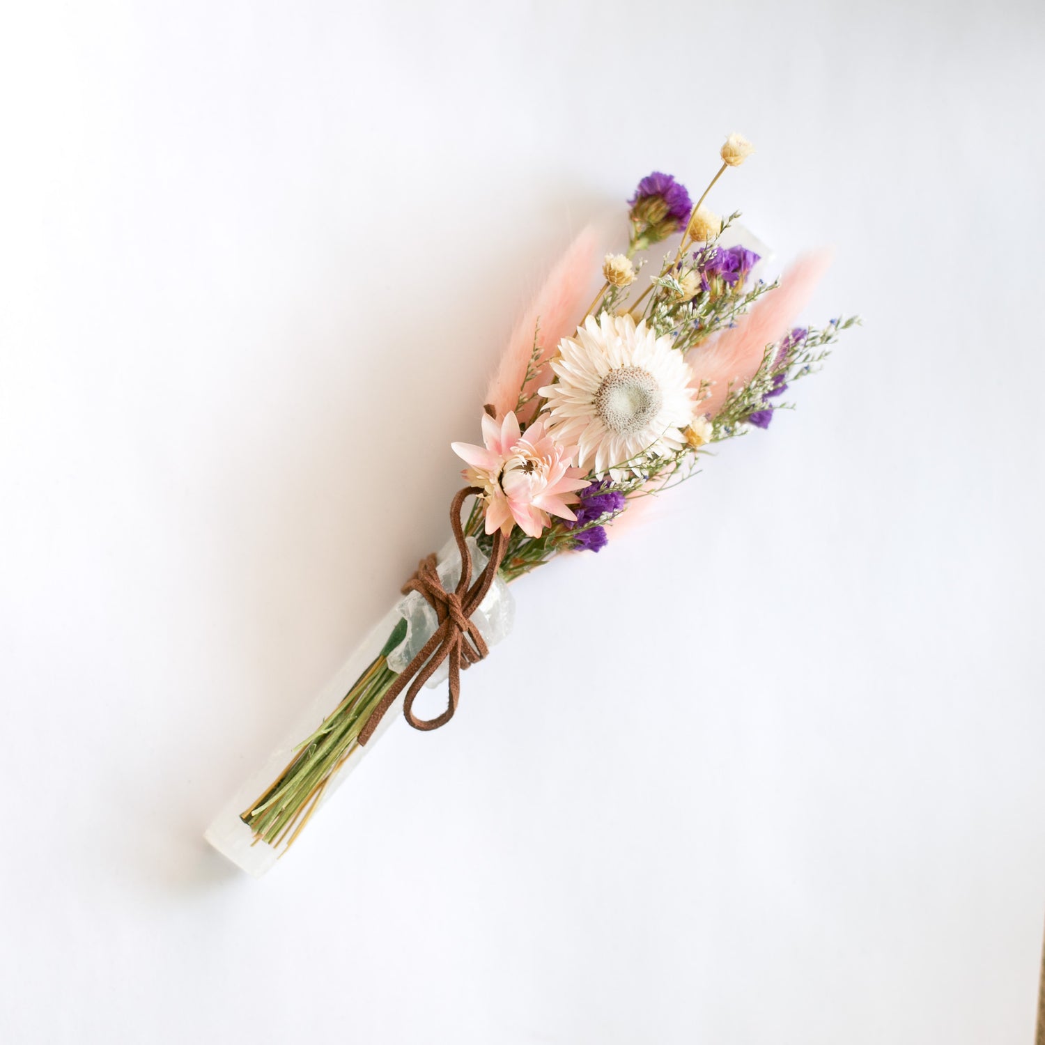 A bundled product flat lay featuring a small dried floral bouquet tied to a stick of selenite and a small quartz crystal on a against a white background.