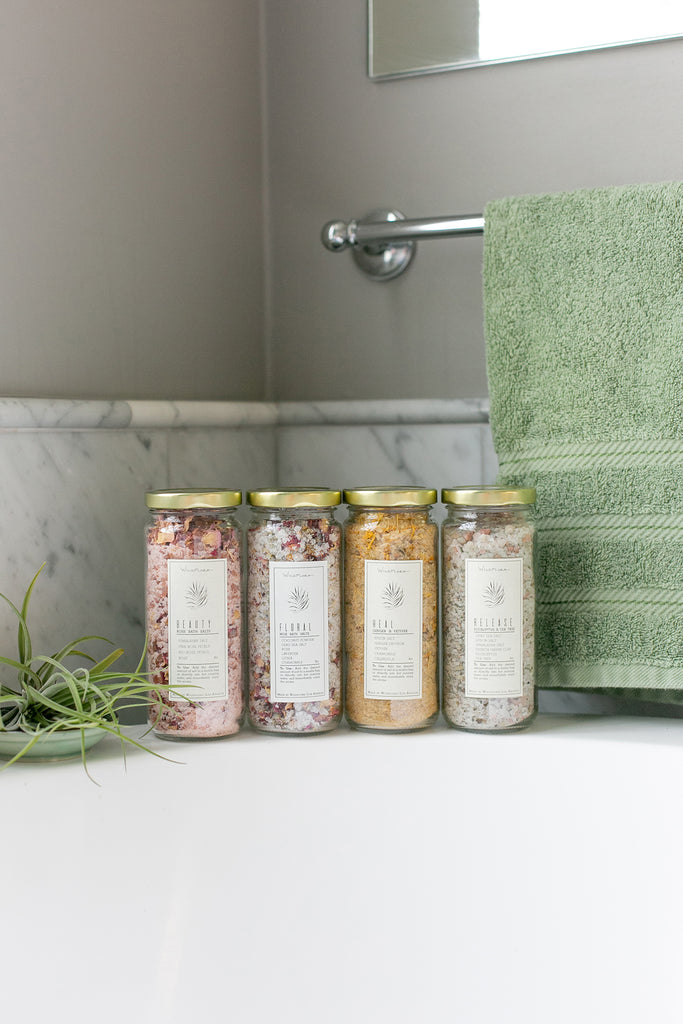 Four jars of bath salts on a bath tub with a green towel and plant in the background.