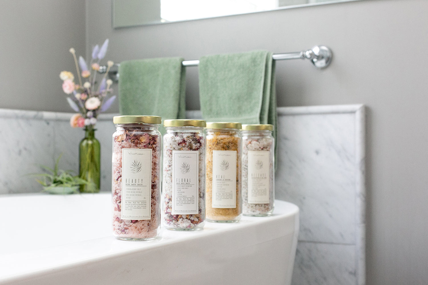 Four jars of bath salts on the edge of a bath tub with sage green towels and dried flowers in the background.