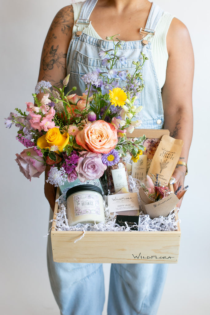 A person in light blue denim overalls holds a wooden gift box filled with a colorful flower bouquet, candles, and packaged WildFlora botanical items. The box is branded WildFlora.