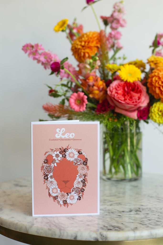 A greeting card on a table with flowers in the background. the card featuring a stylized lion face silhouette in terracotta, framed by a floral mane in white, pink, and dark brown. The print has a peach background with the word 'Leo' and adjectives printed in white at the top.
