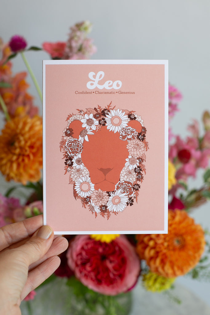 a handing holding A greeting card featuring a stylized lion face silhouette in terracotta, framed by a floral mane in white, pink, and dark brown. The print has a peach background with the word 'Leo' and adjectives printed in white at the top, and is photographed with flowers in the background