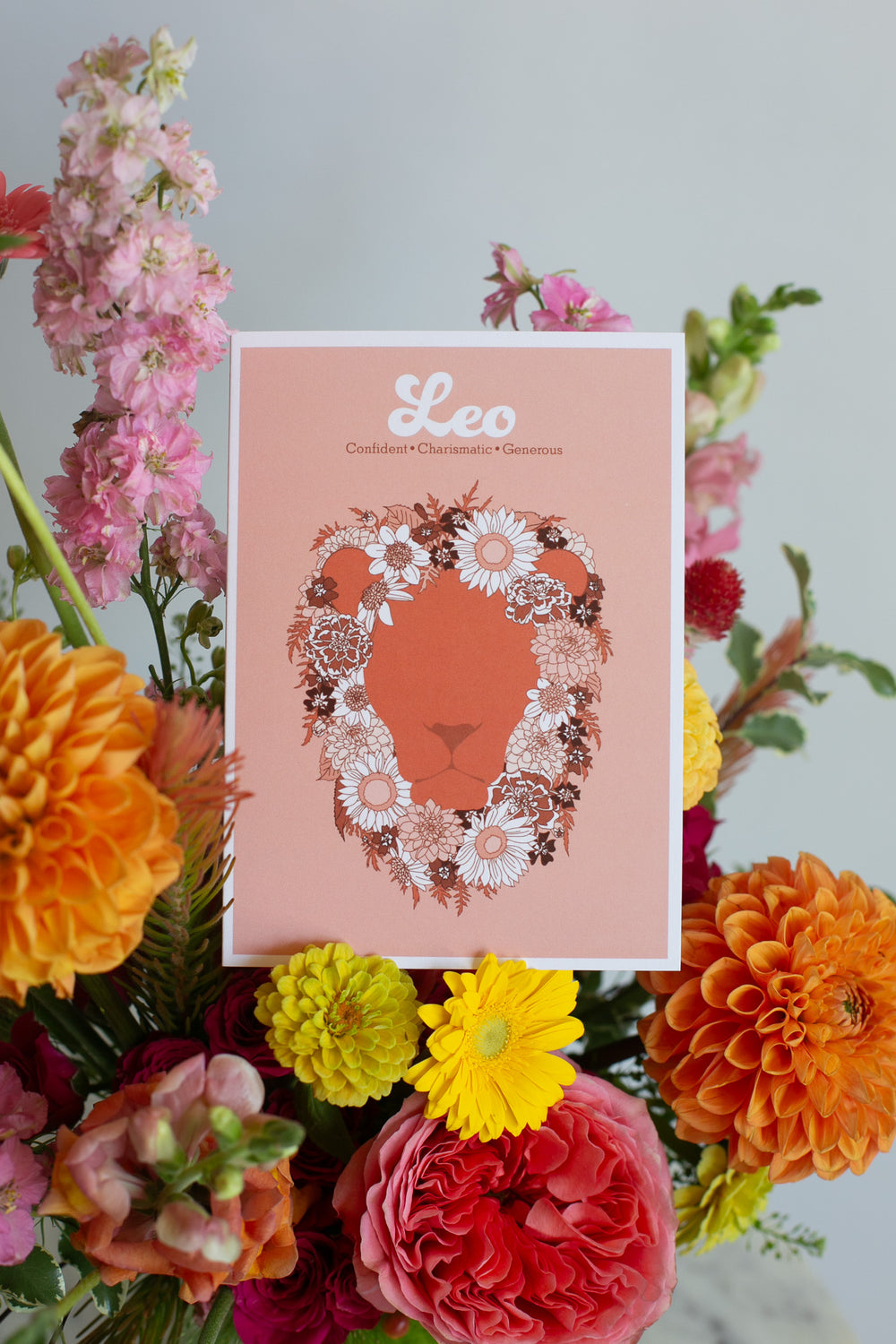 A greeting card featuring a stylized lion face silhouette in terracotta, framed by a floral mane in white, pink, and dark brown. The print has a peach background with the word 'Leo' and adjectives printed in white at the top, and is photographed with flowers in the background