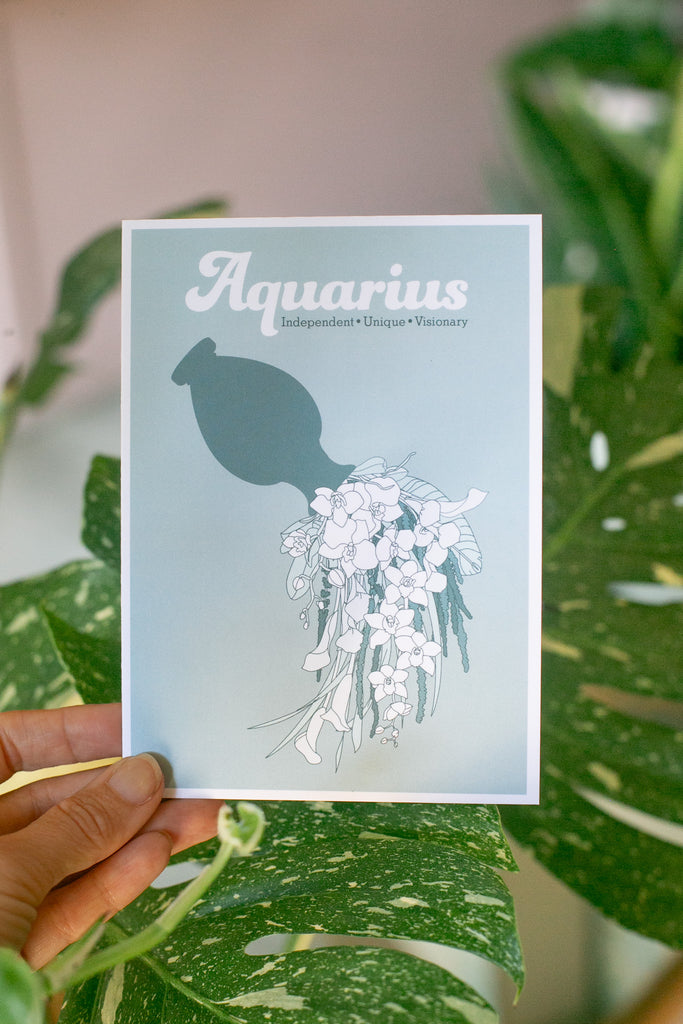 a hand holding A greeting card featuring the word 'Aquarius' in white script above a teal silhouette of a jug pouring illustrated florals on a muted blue-green background, photographed with green leaves in the background.