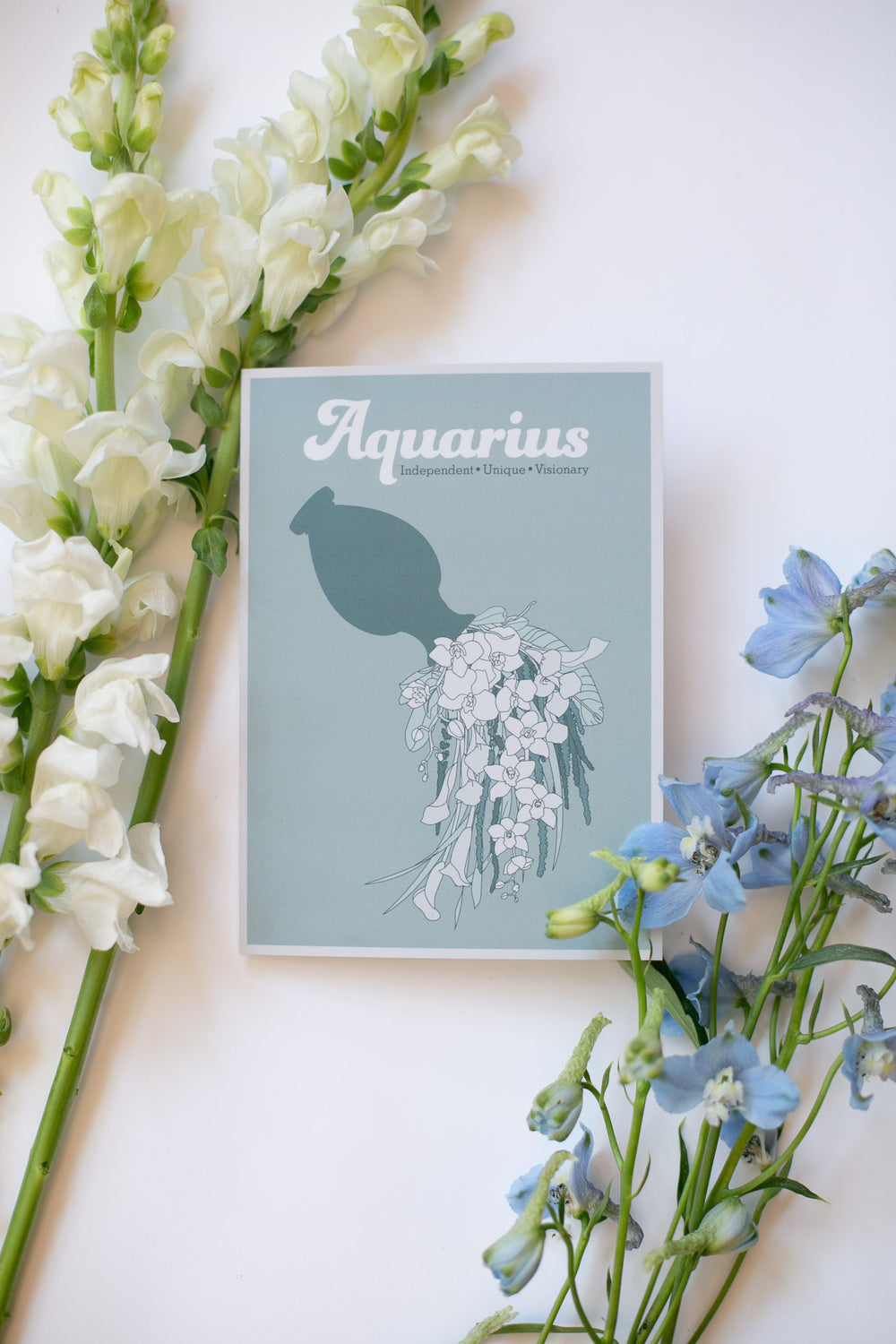 A greeting card featuring the word 'Aquarius' in white script above a teal silhouette of a jug pouring illustrated florals on a muted blue-green background, photographed against a white background with white and blue flowers.