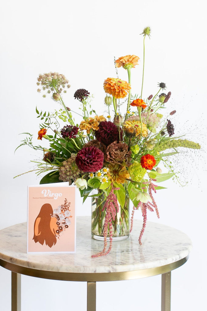 A vibrant wildflower arrangement in a clear glass vase, displayed on a round marble-top side table with a gold metal frame. An orange greeting card with the name 'Virgo' is placed in front of the vase.