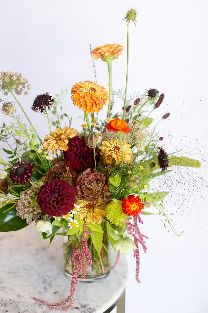 A rustic wildflower bouquet in a clear glass jar vase, featuring orange, burgundy, yellow, and green flowers with delicate pink and purple filler stems, set on a white marble tabletop against a white background.