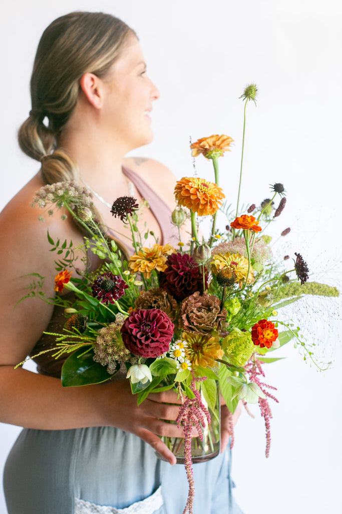 A woman in a dusty pink top and light blue pants holds a vibrant wildflower bouquet in a clear glass vase against a plain white background.