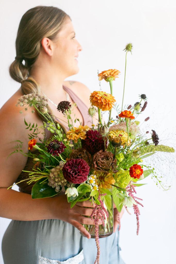 A woman in a dusty pink top and light blue pants holds a vibrant wildflower bouquet in a clear glass vase against a plain white background.