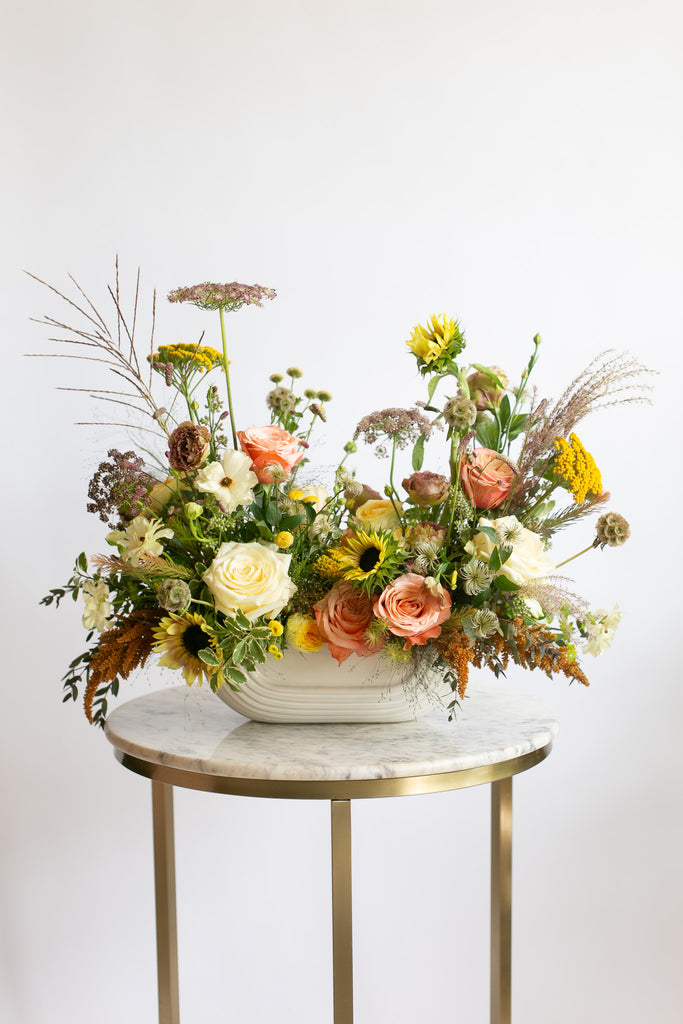 A rustic dried flower arrangement in a white ribbed ceramic bowl, displayed on a round marble-top side table with gold legs against a clean white background.