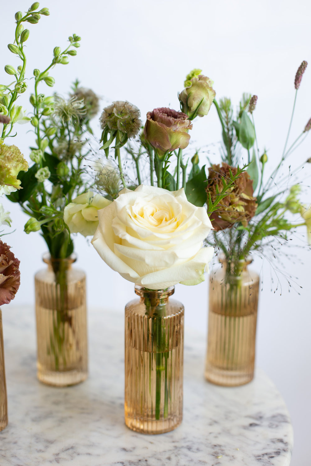 A styled floral arrangement featuring a prominent cream rose in a ribbed amber glass bottle vase, surrounded by other dried and fresh flowers in matching glass vases on a marble surface.