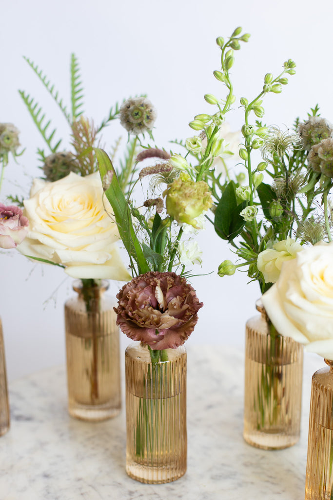 A styled floral arrangement featuring cream roses, green foliage, and a dusty burgundy carnation, presented in ribbed amber glass vases on a light marble surface against a white background.