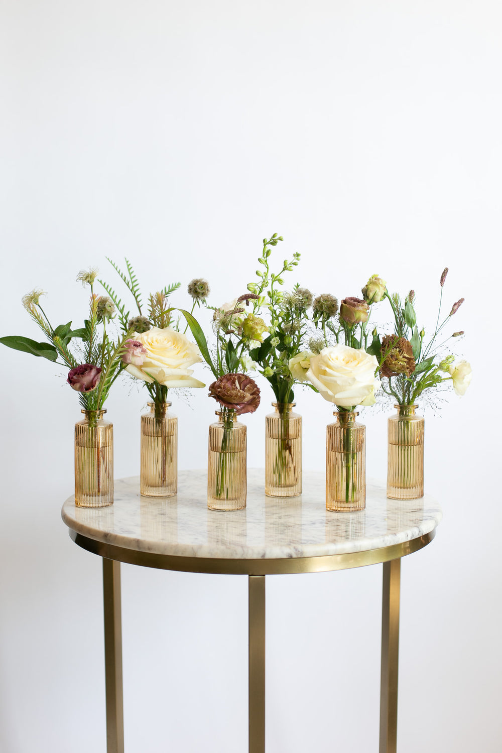 Six small amber glass vases filled with mixed floral arrangements in cream, burgundy, and green tones, displayed on a round marble-top side table with a gold metal frame against a white wall.