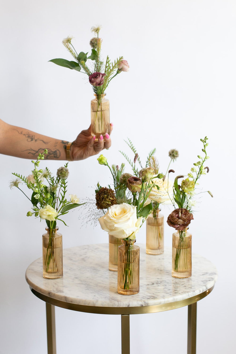 A hand holds a small amber glass vase with a floral arrangement above a round marble-top table with gold legs. Five similar vases with mixed flowers are arranged on the table against a plain white wall.
