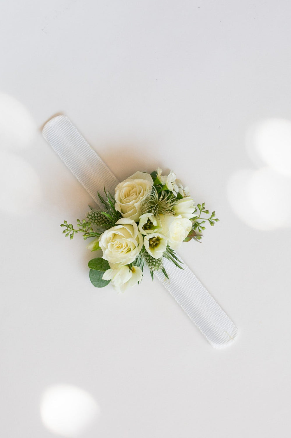 A small floral wrist corsage with white roses and green foliage, attached to a clear plastic tube, shown from a top-down view on a white background with soft bokeh light spots.