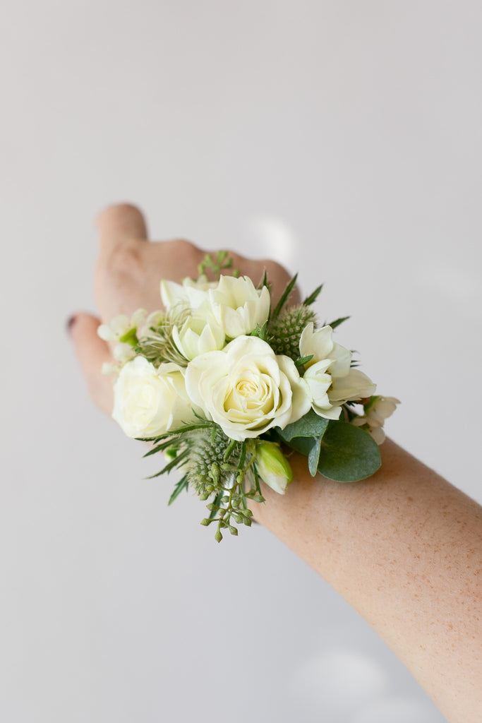 A white rose wrist corsage with green foliage and white lisianthus flowers, worn on a person's wrist against a light gray background.