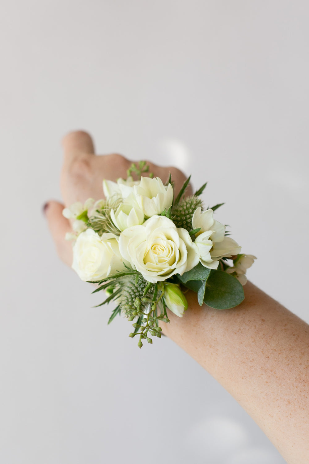 A white rose wrist corsage with green foliage and white lisianthus flowers, worn on a person's wrist against a light gray background.