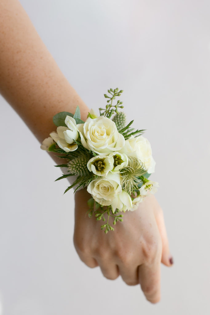 A wrist corsage featuring white roses, white lilies, and green foliage, worn on a person's wrist against a plain light gray background.