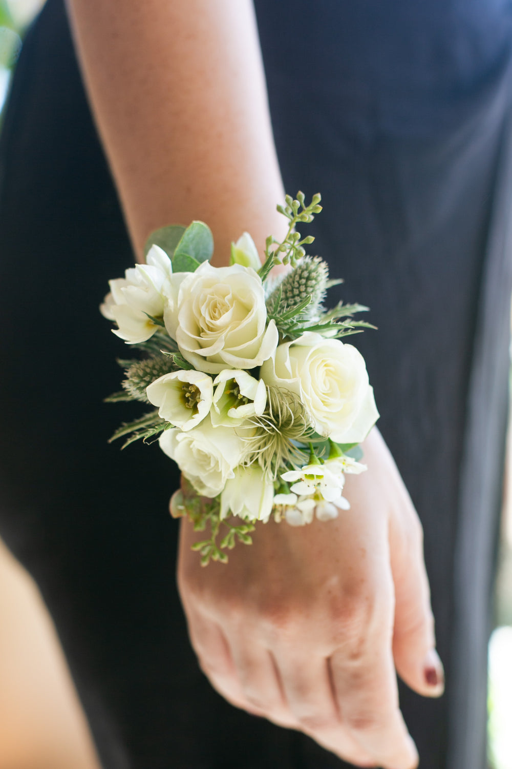 A close-up of a wrist corsage featuring white roses, lisianthus, and green foliage worn on a person's wrist, with a black dress in the background.