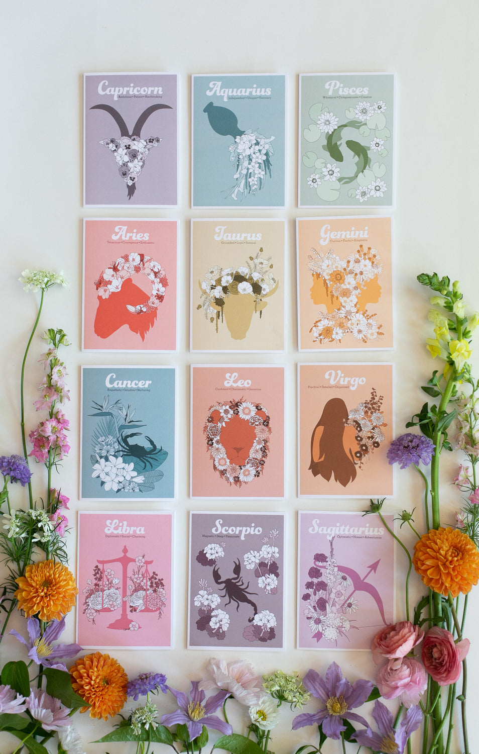 A set of twelve pastel zodiac greeting cards arranged in a 3x4 grid on a white background, surrounded by colorful flowers. The prints feature minimalist illustrations of zodiac symbols and names: Capricorn, Aquarius, Pisces, Taurus, Leo, Gemini, Cancer, Scorpio, Virgo, Libra, Sagittarius, and Scorpio.