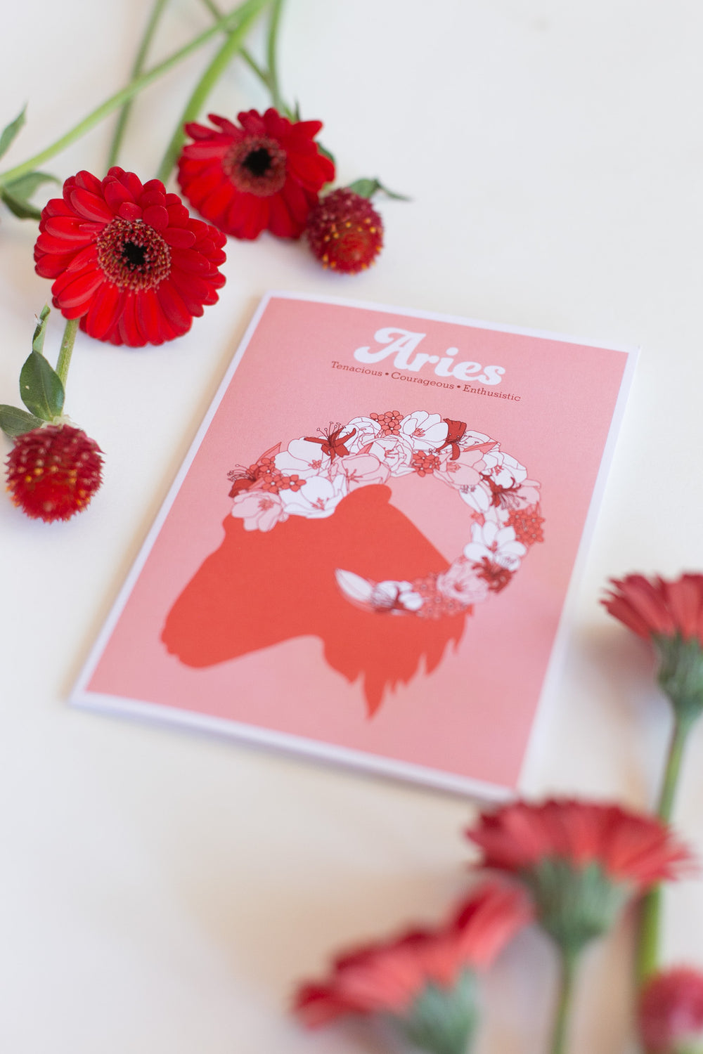 A greeting card featuring a stylized red Aries zodiac silhouette with a white floral crown and the word 'Aries' at the top, styled on a white surface with red flowers around it.