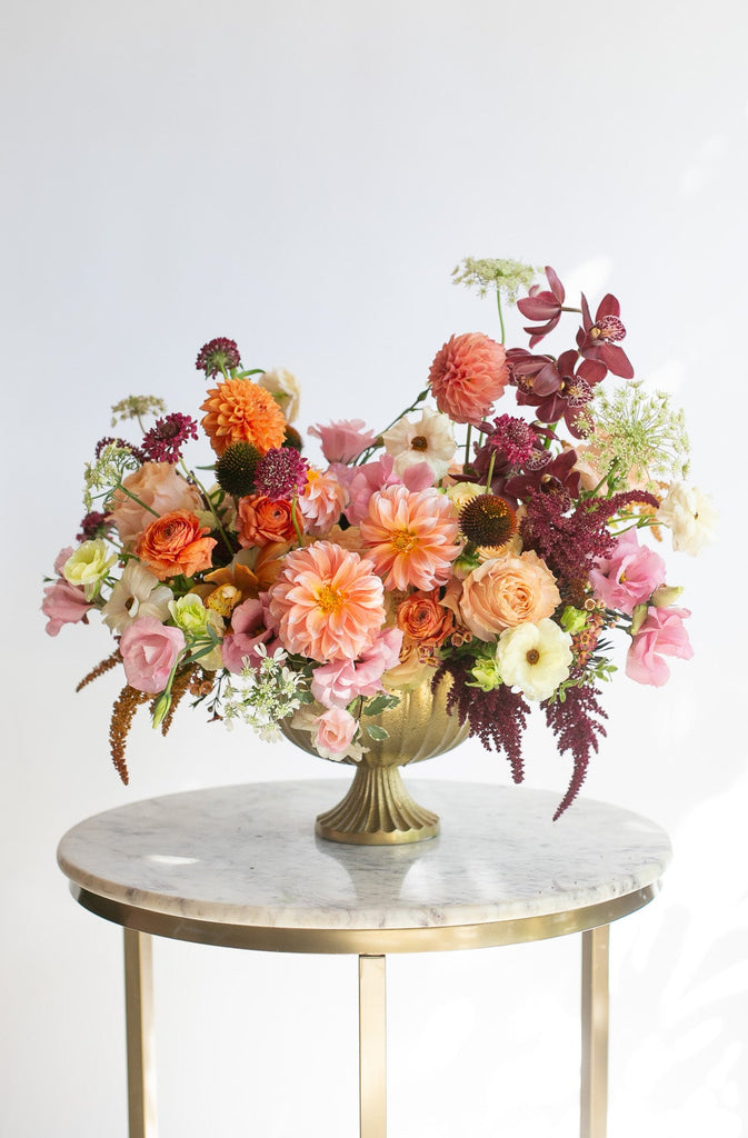 A vibrant, warm-toned floral arrangement with peachy pink dahlia & lisianthus, maroon orchids, white and orange ranunculus, and tan and dark purple amaranths in a gold urn vase, displayed on a round marble-top side table with gold legs against a clean white background.