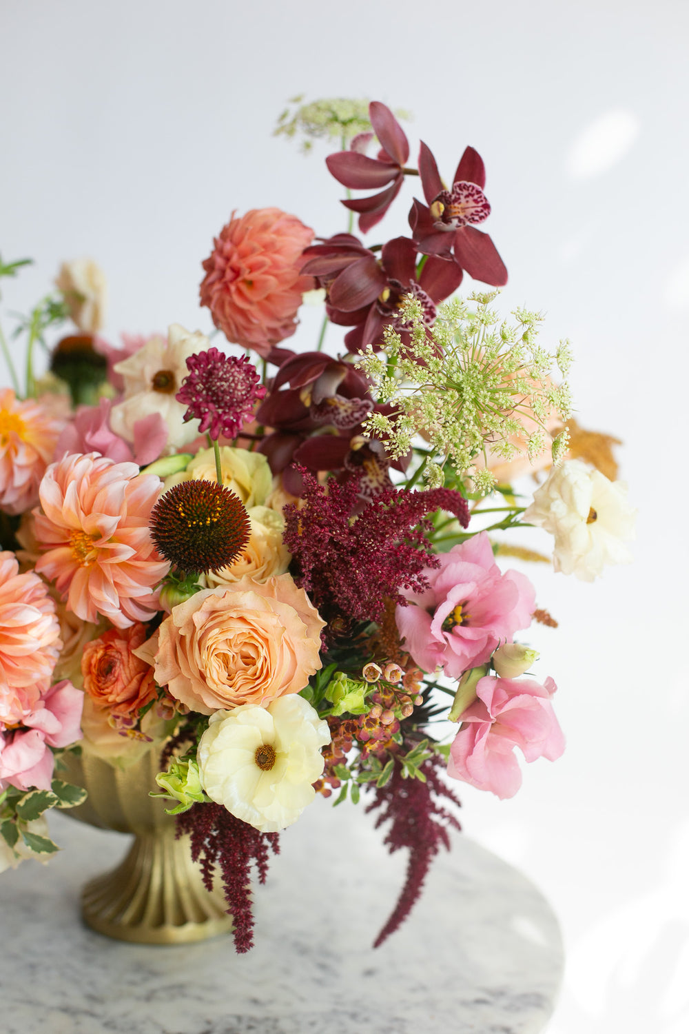 A vibrant, warm-toned floral arrangement with peachy pink dahlia & lisianthus, maroon orchids, white and orange ranunculus, and tan and dark purple amaranths in a gold urn vase, displayed on a round marble-top side table with gold legs against a clean white background.
