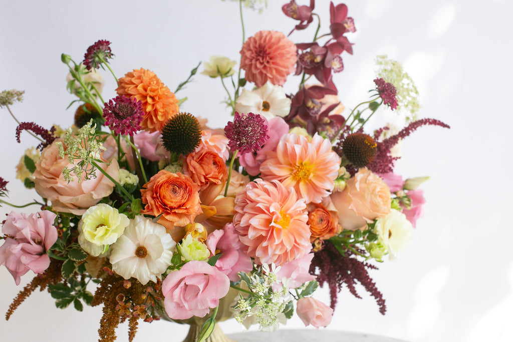 A vibrant, warm-toned floral arrangement with peachy pink dahlia & lisianthus, maroon orchids, white and orange ranunculus, and tan and dark purple amaranths in a gold urn vase, displayed against a clean white background.