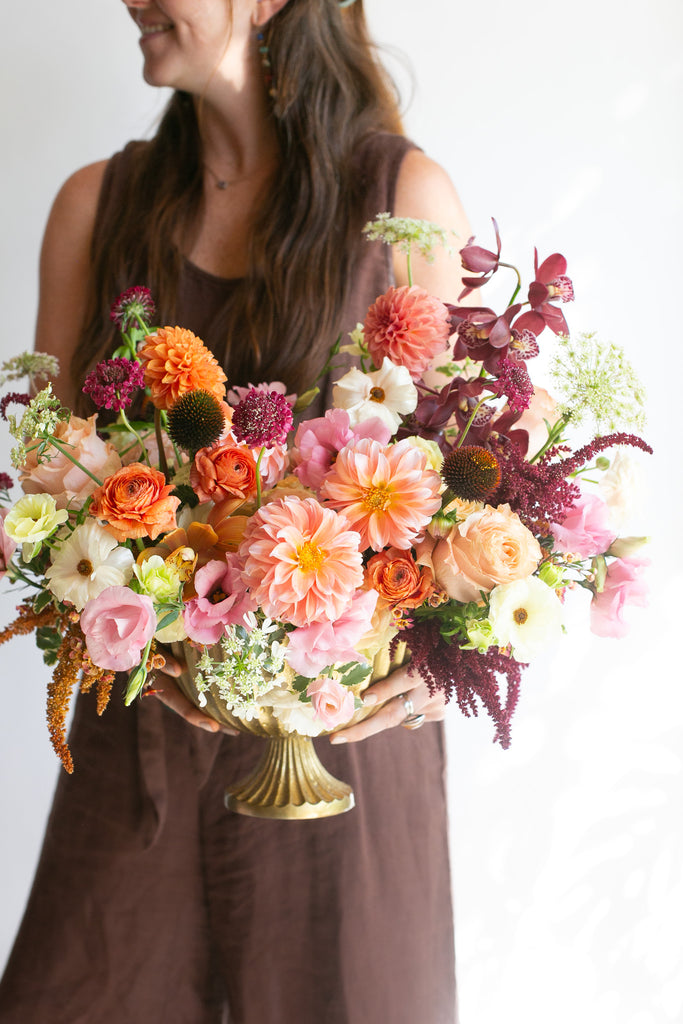 A woman in a brown sleeveless dress holds aA vibrant, warm-toned floral arrangement with peachy pink dahlia & lisianthus, maroon orchids, white and orange ranunculus, and tan and dark purple amaranths in a gold urn vase against a clean white background.