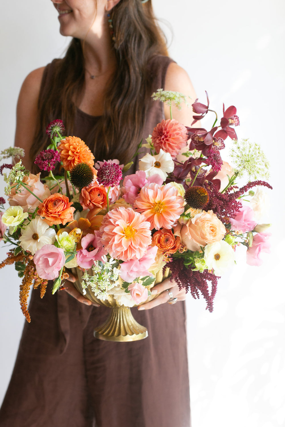 A woman in a brown sleeveless dress holds aA vibrant, warm-toned floral arrangement with peachy pink dahlia & lisianthus, maroon orchids, white and orange ranunculus, and tan and dark purple amaranths in a gold urn vase against a clean white background.