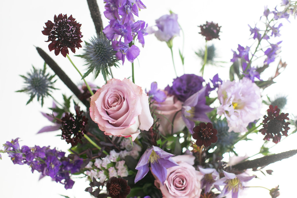 A close-up floral arrangement featuring light pink roses, purple clematis vine, lisianthus, delphinium, dark burgundy scabiosa, blue globe thistle, and green foliage against a bright white background.
