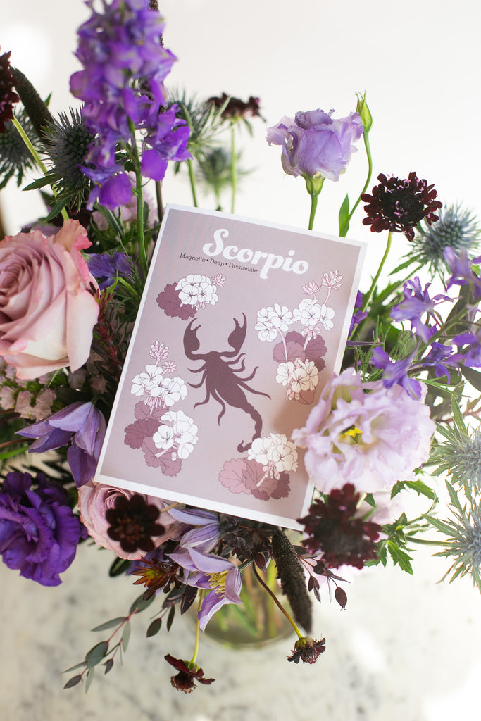 A Scorpio zodiac card with a purple scorpion and floral design, nestled inside a colorful bouquet of purple & pink flowers.