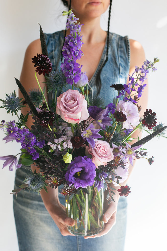 A person in a denim jumpsuit holds a vibrant flower bouquet in a clear glass vase. The bouquet features prominent purple flowers, light pink roses, and green foliage against a plain white background.