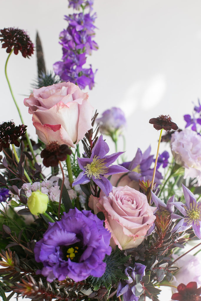 A close-up floral arrangement featuring light pink roses, purple clematis vine, lisianthus, delphinium, dark burgundy scabiosa, blue globe thistle, and green foliage against a bright white background.