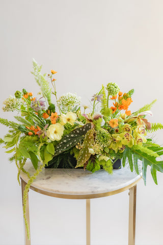 A flower arrangement in a matte, long & low black vase in front of a white backdrop on a marble table. It's filled with orange, white, purple, but mostly green and and chartreuse-colored flowers and foliage, including sword fern, fritillaria, alium, pitcher plant, anemone, ornamental grass, polka dot begonia, asparagus fern, star of bethlehem, clematis, and cymbidium orchid. The vibes are very Hobbiton.